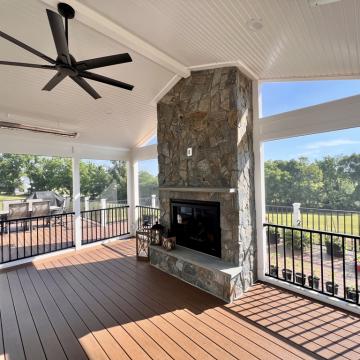 Screened open-air porch with stone fireplace, ceiling fan, and deck views