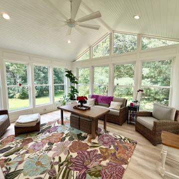 Bright enclosed sunroom with vaulted beadboard ceiling, large windows, wicker seating, floral area rug, ceiling fan, and lush garden views.